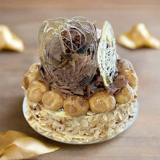 A decadent layered cake with pastry and custard layers, wrapped in roasted slivered almonds, and topped with custard filled, toffee dipped profiteroles, and chocolate shavings.